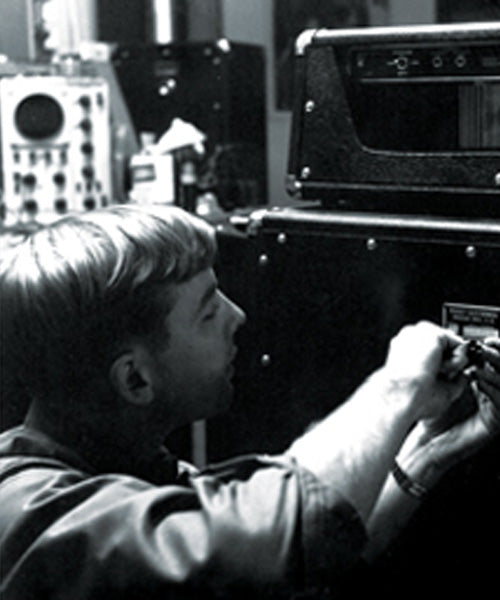 Black and white photo of a man in profile working at a control panel or equipment in a technical room, with large electronic devices in the background
